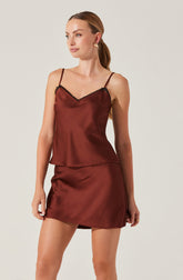 A woman is wearing a sleeveless satin camisole top with thin spaghetti straps and a V neckline accented with black lace trim. She pairs it with a matching satin mini skirt. The outfit is in a rich, reddish-brown color.